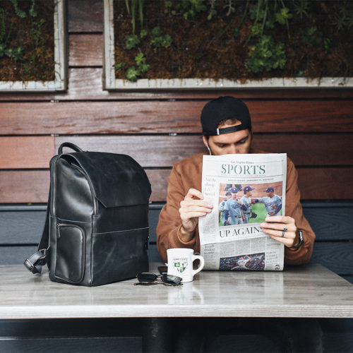 A man reading the sports section of the newspaper in a coffee shop with his black leather work backpack sitting beside him