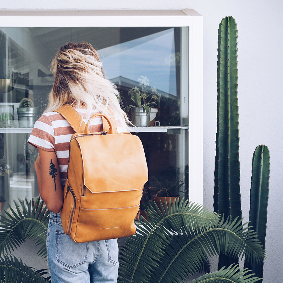 A blonde woman wearing a caramel-colored leather backpack facing backward next to a cactus