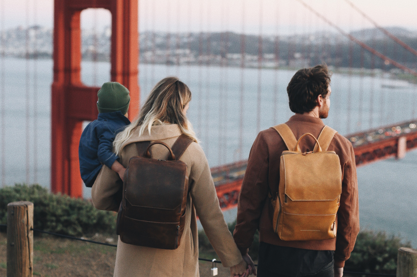 A happy couple wearing genuine leather backpacks while carrying their baby boy and looking at the Golden Gate Bridge